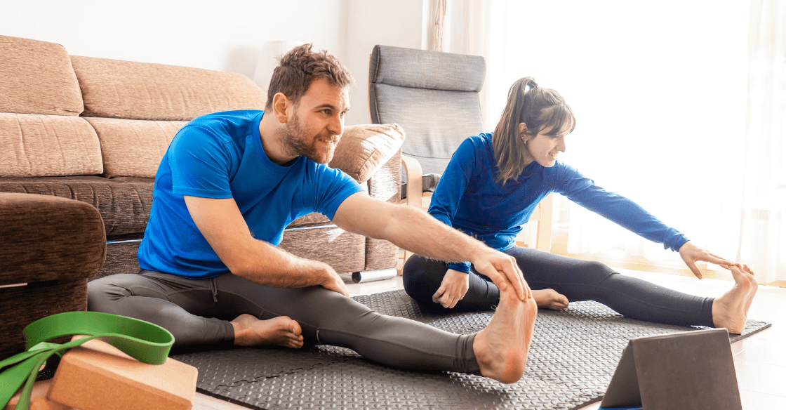 Couple exercising in living room following video from tablet woman and man stretched out on exercise mat with tablet and yoga blocks